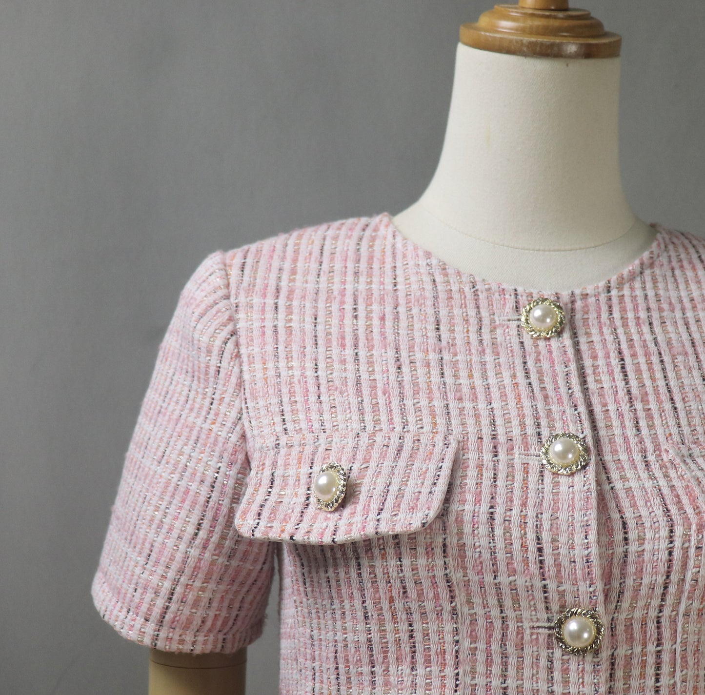 Close-up of pink tweed fabric showing texture, detailed stitching, and decorative gold-tone buttons on the blazer for an elegant finish.