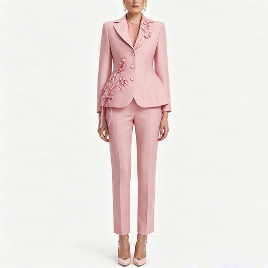A model wears a light pink tailored suit consisting of a blazer with floral applique and matching slim trousers, paired with pink high heels against a plain white background.