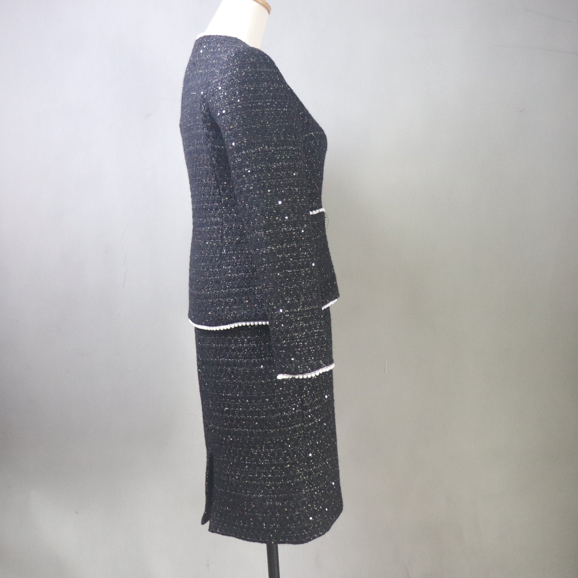A side view of a custom-made black tweed skirt suit for women on a white mannequin. The two-piece set features a tailored V-neck jacket with shimmering silver sequins, white pearl-beaded trim, and a white floral camellia brooch. The matching pencil skirt is knee-length, highlighting the textured, sparkly tweed fabric against a neutral gray background.