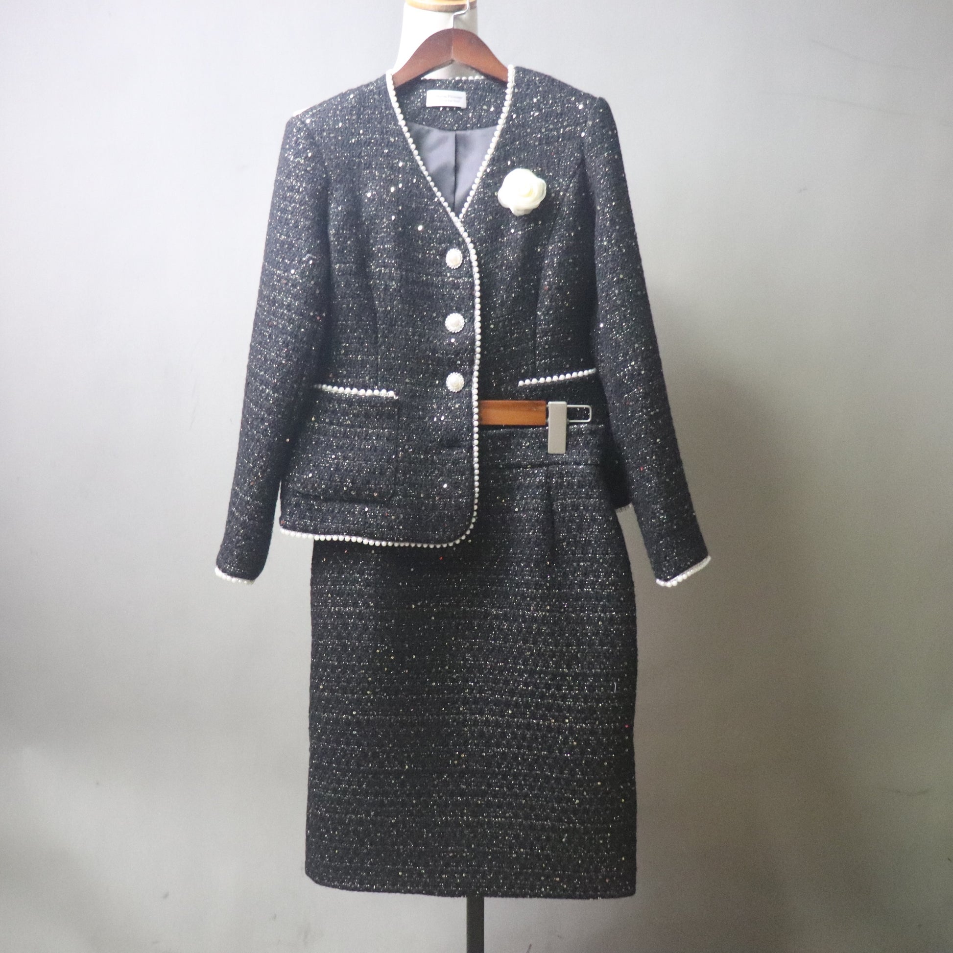 A front view of a custom-made black tweed skirt suit for women on a white mannequin. The two-piece set features a tailored V-neck jacket with shimmering silver sequins, white pearl-beaded trim, and a white floral camellia brooch. The matching pencil skirt is knee-length, highlighting the textured, sparkly tweed fabric against a neutral gray background.