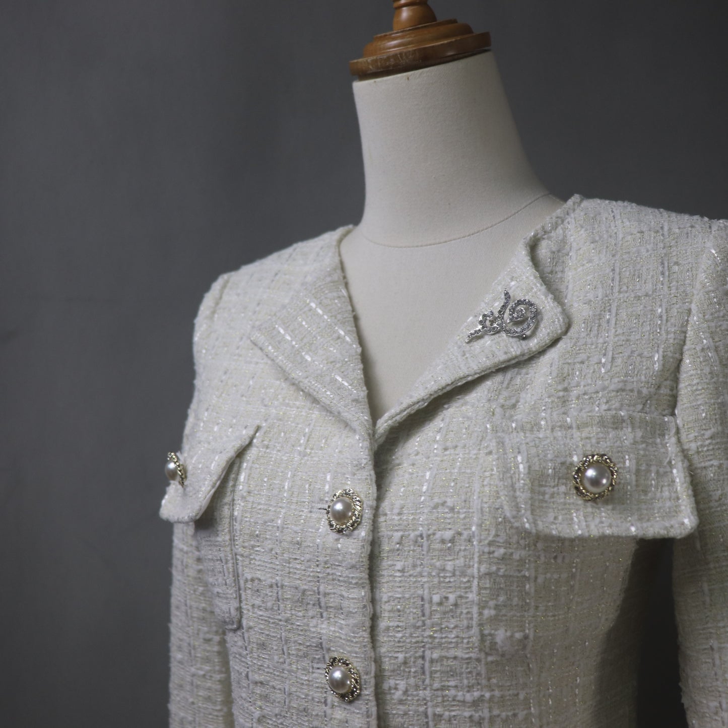 Close-up of the shoulder and chest area of a white boucle tweed jacket on a mannequin. Features include large pearl buttons, buttoned flap pockets, a lapel collar, and a decorative rhinestone brooch pinned to the collar.