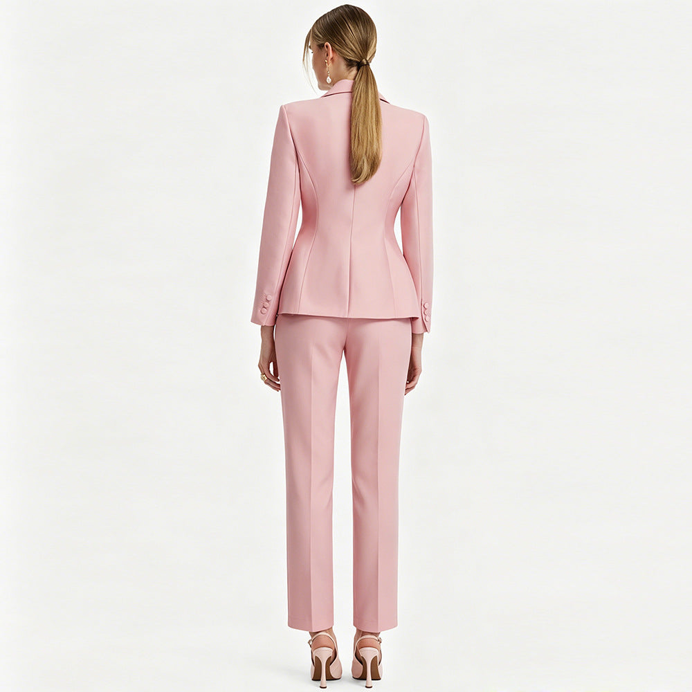A woman shown from the back wearing a light pink tailored suit with a structured blazer and straight-leg trousers, paired with metallic high heels against a plain light background.