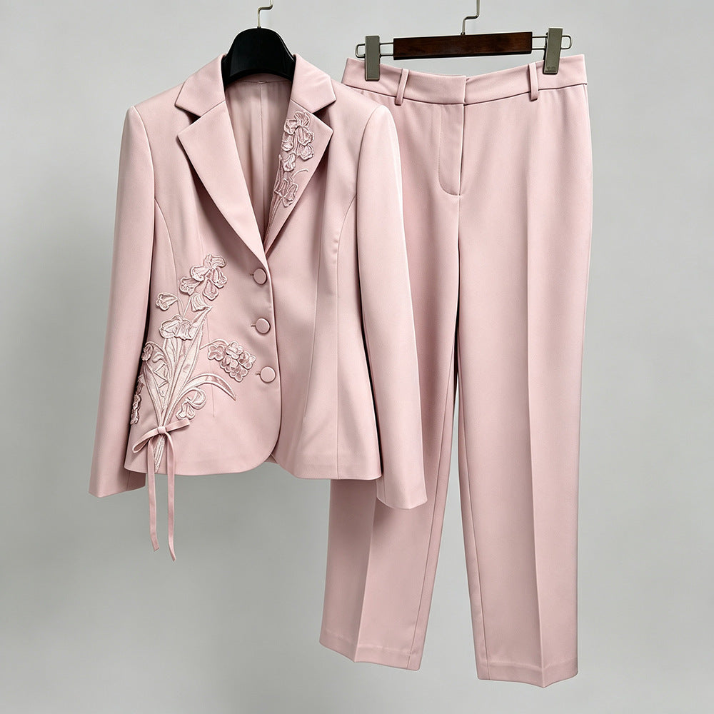 A two-piece women's suit set in light pink, featuring a single-breasted blazer with floral embroidery on the lapel and sleeve, and matching straight-leg trousers, displayed on hangers against a neutral gray background.
