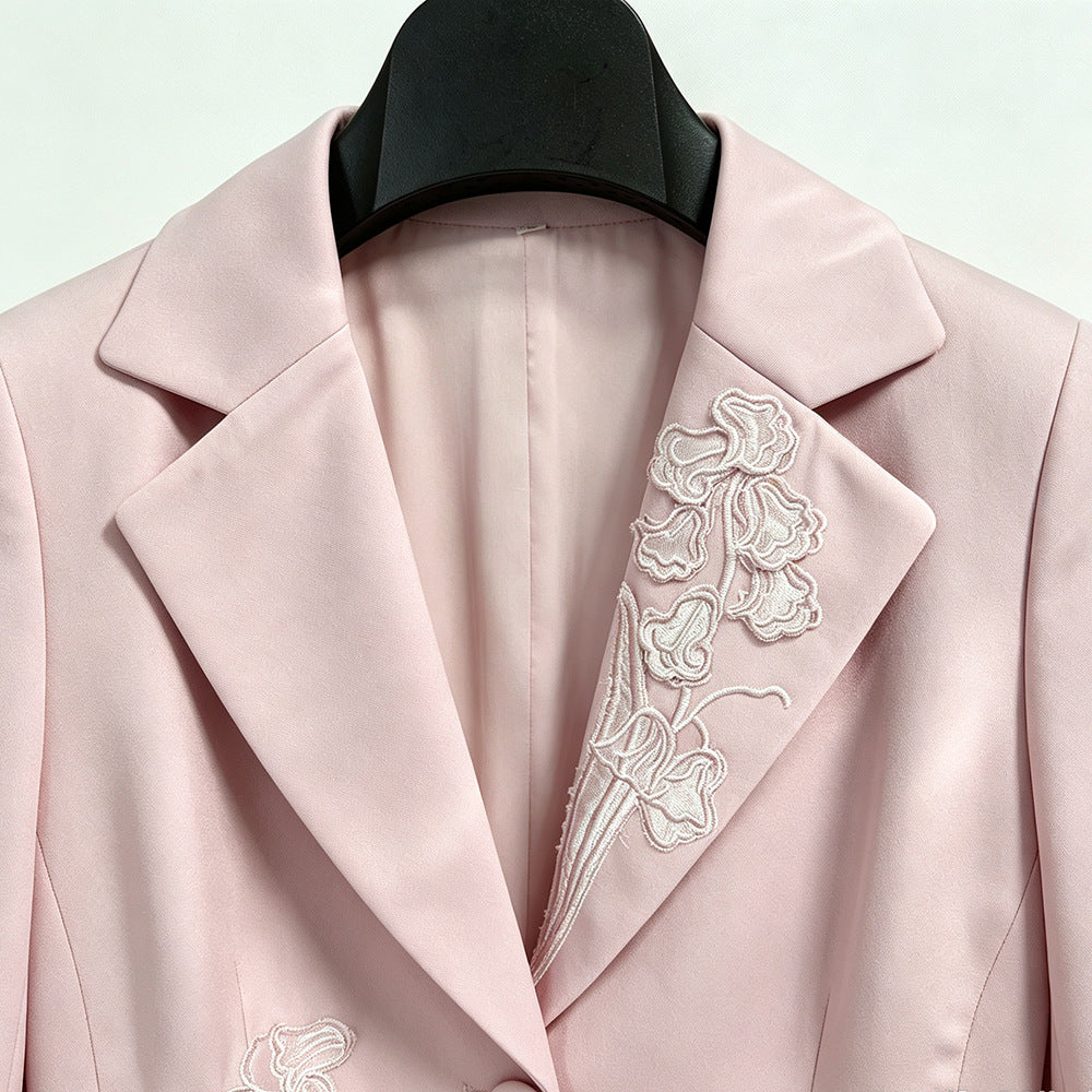 A close-up of a light pink blazer with white floral embroidery on the lapel, displayed on a black hanger against a light background.