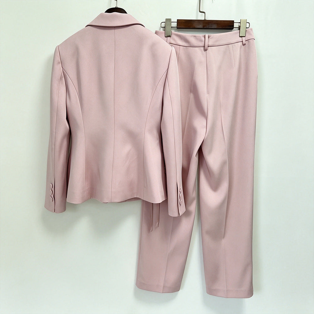 A two-piece women's suit set in dusty pink, featuring a tailored blazer and matching trousers, displayed on hangers against a plain off-white wall.