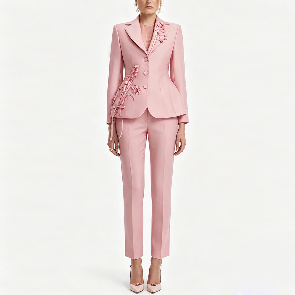 A model wears a light pink tailored suit consisting of a blazer with floral applique and matching slim trousers, paired with pink high heels against a plain white background.