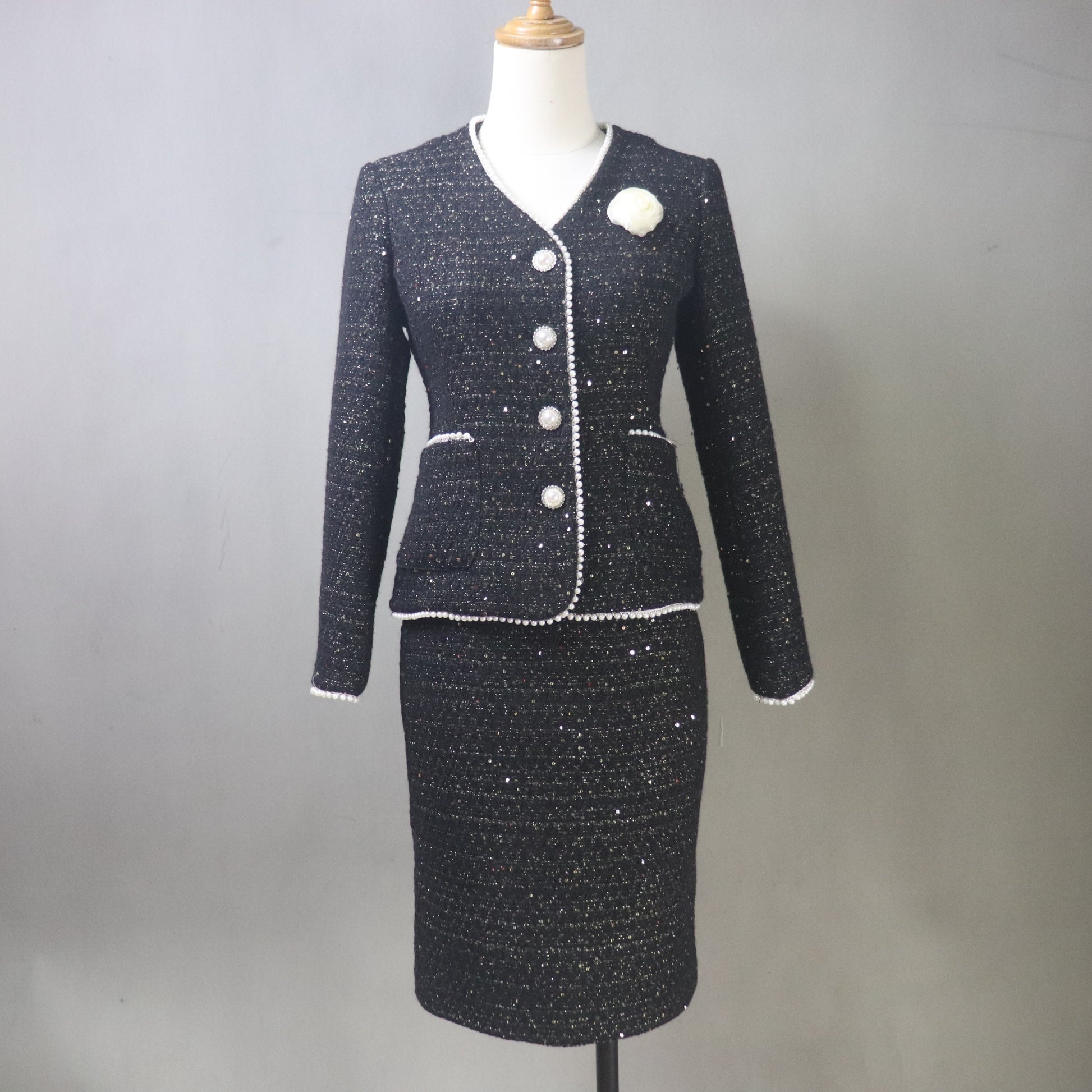 A front view of a custom-made black tweed skirt suit for women on a white mannequin. The two-piece set features a tailored V-neck jacket with shimmering silver sequins, white pearl-beaded trim, and a white floral camellia brooch. The matching pencil skirt is knee-length, highlighting the textured, sparkly tweed fabric against a neutral gray background.