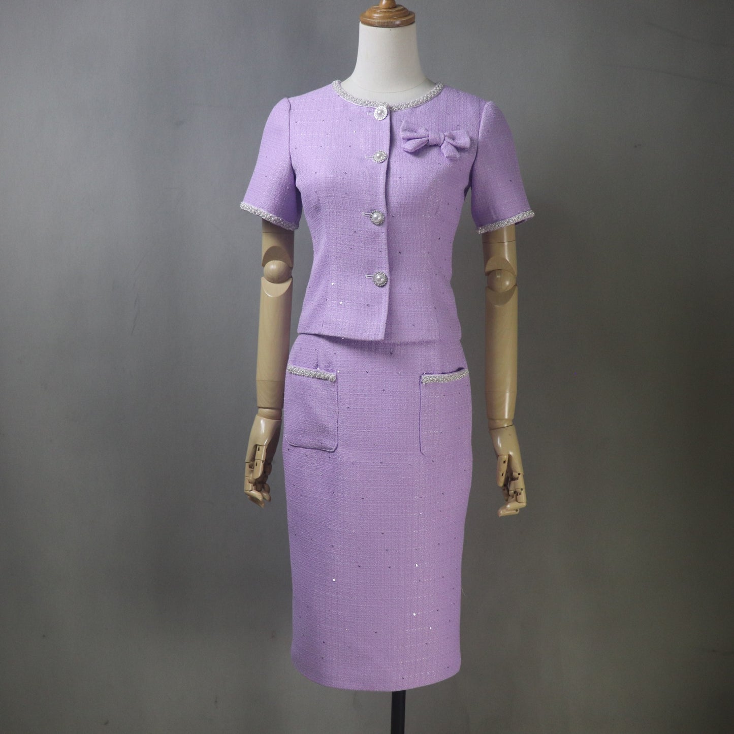 Custom-made lilac tweed suit for women. Elegant two-piece set featuring a short-sleeve, jacket with bow and sparkle buttons, paired with a matching knee-length pencil skirt with trimmed pockets. Made-to-measure tailoring, perfect for Mother of the Bride, professional business wear, or formal occasions.