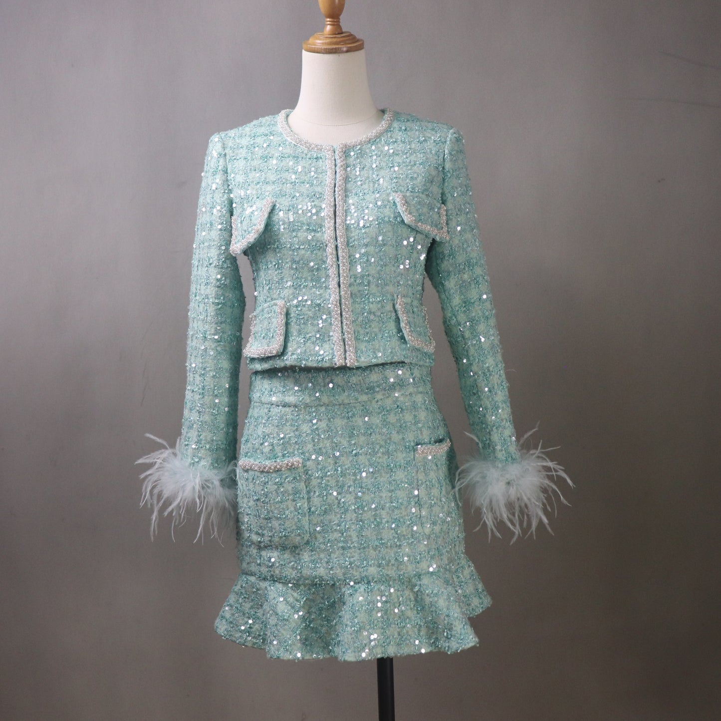 Custom aqua sequin tweed skirt suit for women featuring a fitted jacket with feather cuffs, matching ruffled skirt, and decorative trim displayed on mannequin.