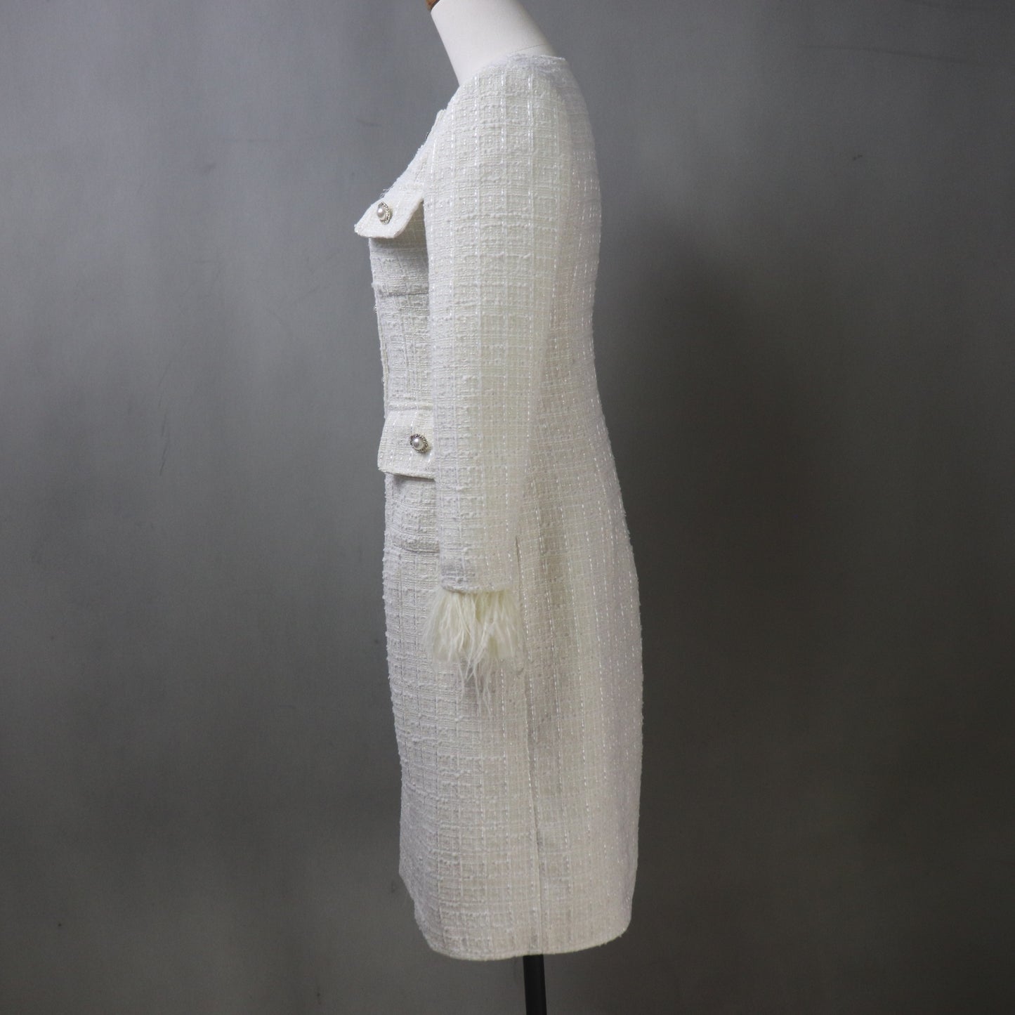Side view of the custom white boucle tweed coat displayed on a mannequin, highlighting the fitted silhouette, textured fabric weave, and the white feather trim on the sleeve cuff.
