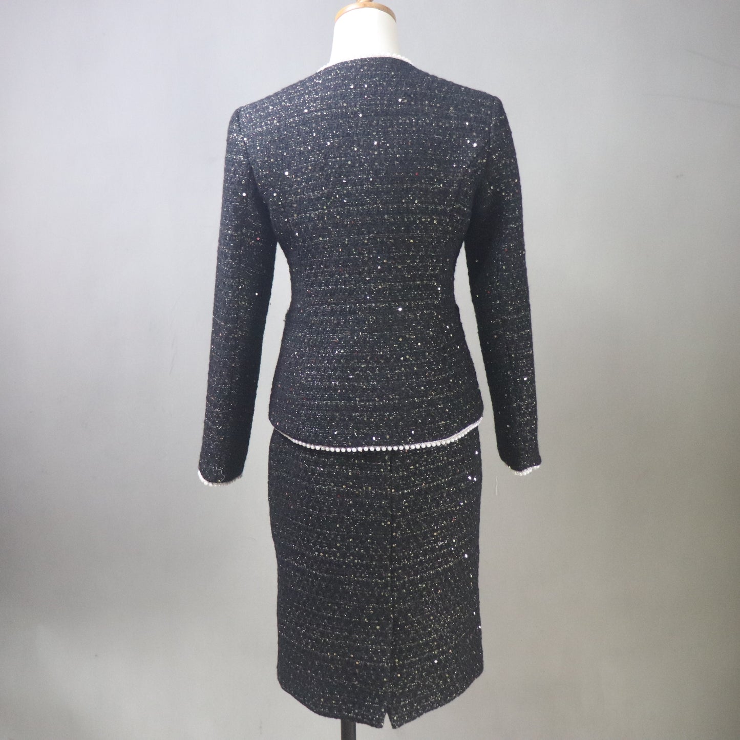 A back view of a custom-made black tweed skirt suit for women on a white mannequin. The two-piece set features a tailored V-neck jacket with shimmering silver sequins, white pearl-beaded trim, and a white floral camellia brooch. The matching pencil skirt is knee-length, highlighting the textured, sparkly tweed fabric against a neutral gray background.