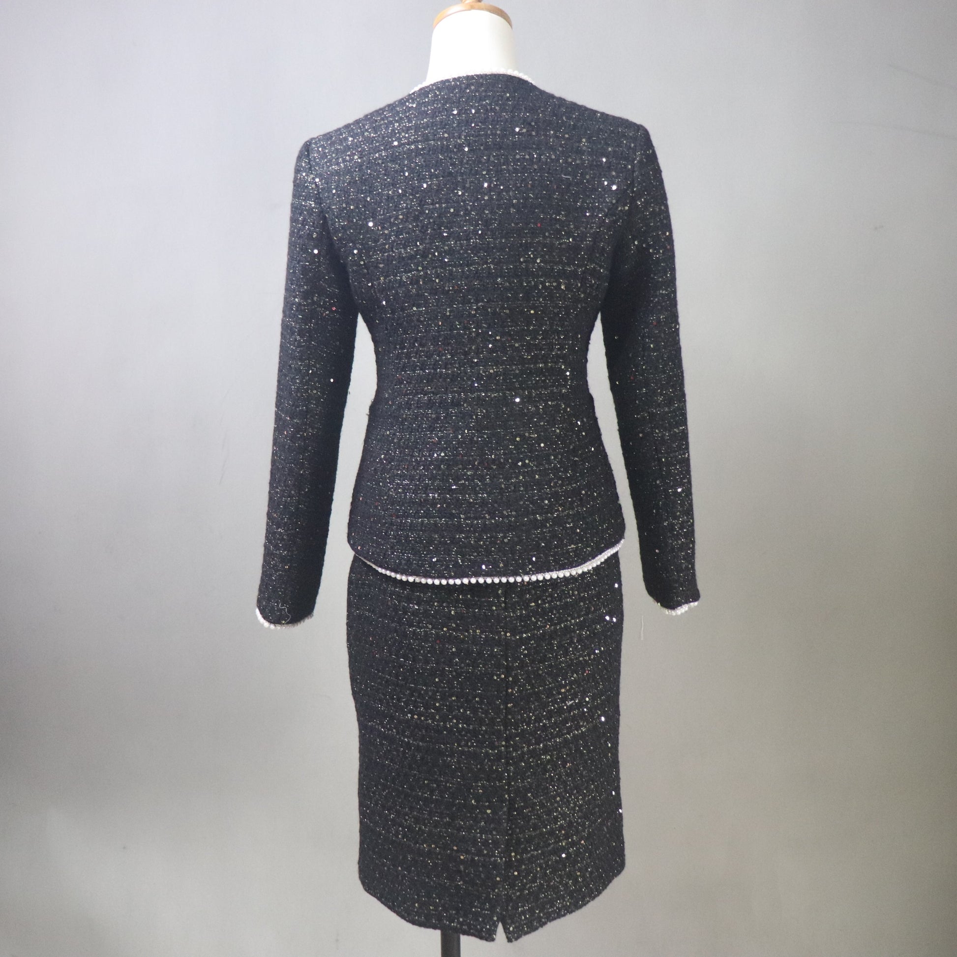 A back view of a custom-made black tweed skirt suit for women on a white mannequin. The two-piece set features a tailored V-neck jacket with shimmering silver sequins, white pearl-beaded trim, and a white floral camellia brooch. The matching pencil skirt is knee-length, highlighting the textured, sparkly tweed fabric against a neutral gray background.