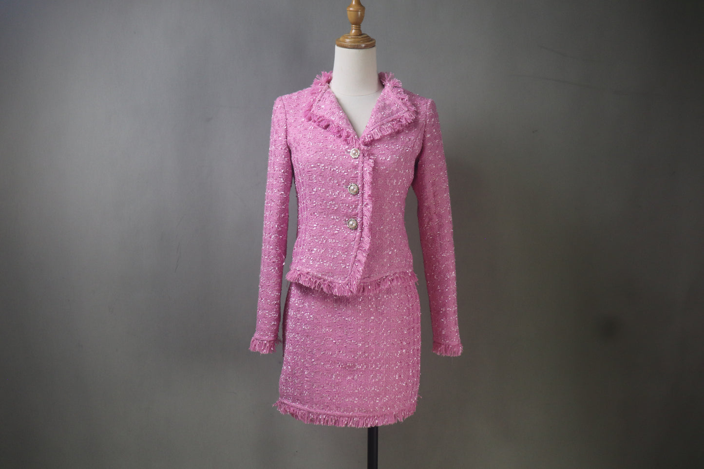 A custom-made, bright pink tweed skirt suit on a mannequin. The suit features a tailored jacket with frayed fringe trim and a matching skirt that can be worn at mini or midi length. High-quality bespoke women's fashion.