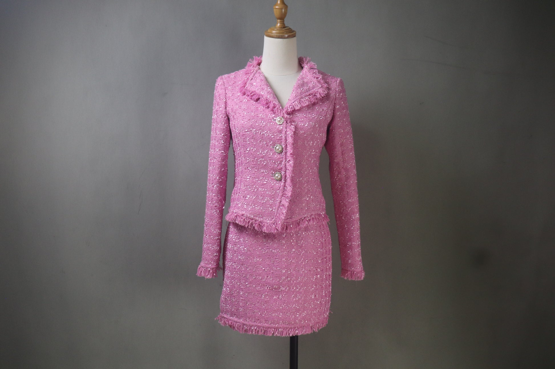 A custom-made, bright pink tweed skirt suit on a mannequin. The suit features a tailored jacket with frayed fringe trim and a matching skirt that can be worn at mini or midi length. High-quality bespoke women's fashion.